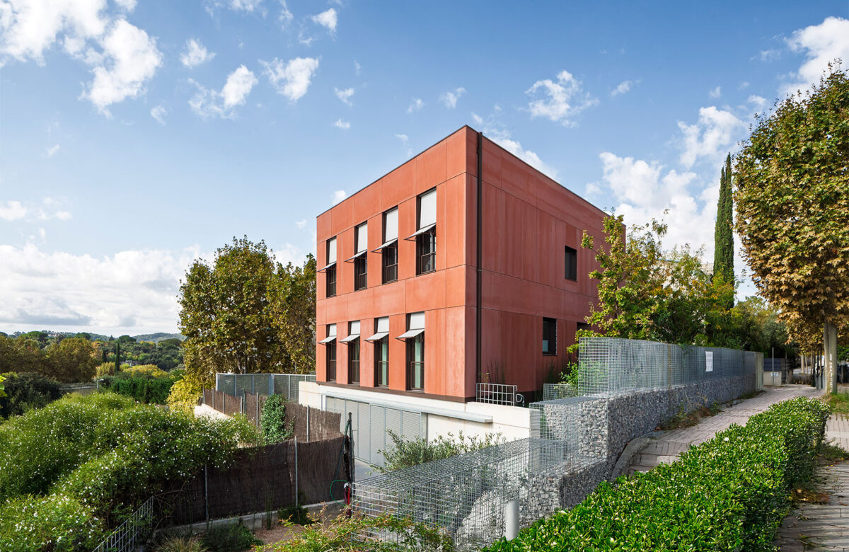 La imatge presenta una construcció de casa passiva que destaca per la seva arquitectura sostenible i eficiència energètica, característiques clau del disseny Passivhaus. L'edifici, de dues plantes, es caracteritza per una façana terracota i finestres grans amb persianes obertes, que combinades amb l'integració de vegetació al voltant del recinte, aporten un aspecte natural i sostenible. La construcció casa passiva es complementa amb elements de disseny que minimitzen el consum d'energia, fent d'aquesta casa un exemple de construcció casa sostenible en Sant Cugat, que combina modernitat i eficiència energètica per a una vida més sostenible.