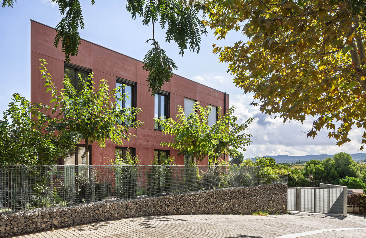 La imatge mostra una casa passiva sostenible amb disseny Passivhaus, caracteritzada per la seva façana terracota i finestres grans amb persianes obertes, que combinades amb l'integració de vegetació al voltant i una tanca metàl·lica, aporten un aspecte modern i sostenible que s'adapta perfectament al seu entorn natural a Sant Cugat.
