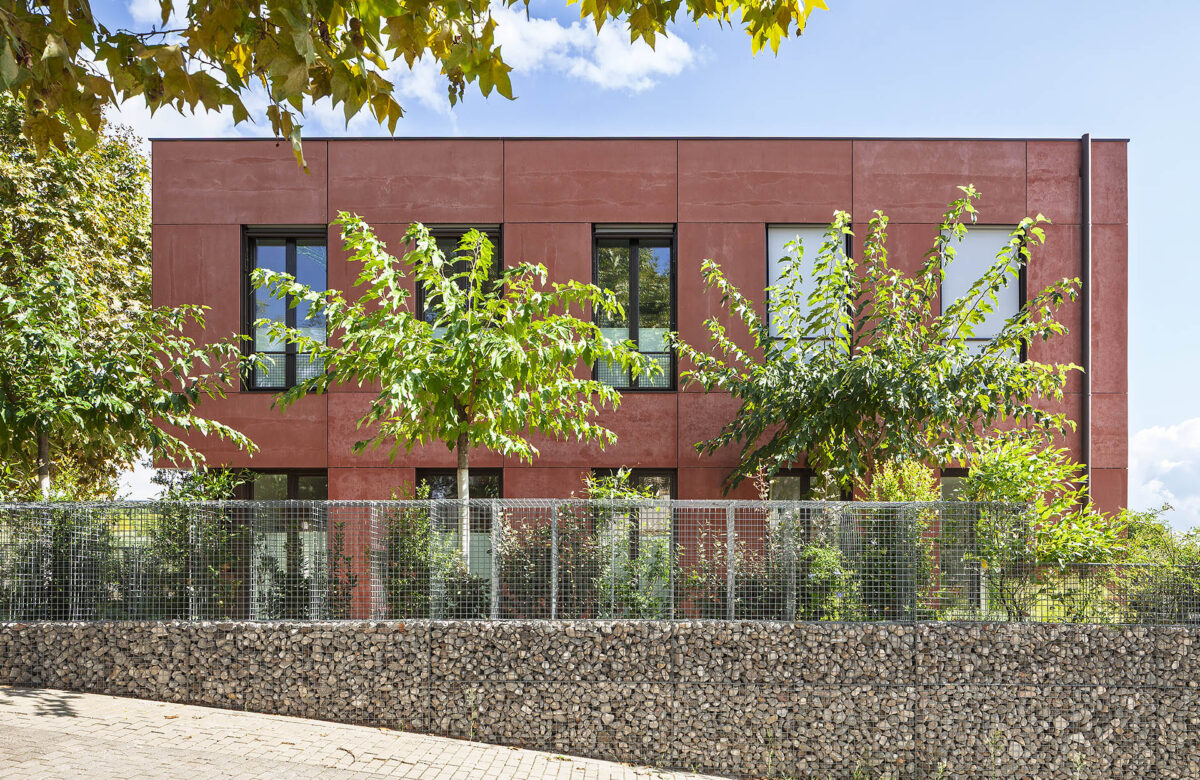 La fotografía presenta una casa pasiva sostenible, diseñada con eficiencia energética en mente, caracterizada por su fachada de color terracota y ventanas grandes que ofrecen luz natural y ventilación adecuada. El edificio, situado en un ambiente verde y tranquilo, está rodeado por una valla de piedra y malla metálica, combinados con vegetación que aporta un aspecto natural y sostenible. La construcción de la casa pasiva en Sant Cugat es un ejemplo de diseño que minimiza el consumo de energía para la calefacción y enfriarlo, consiguiendo el estándar en el diseño de casas sostenibles.