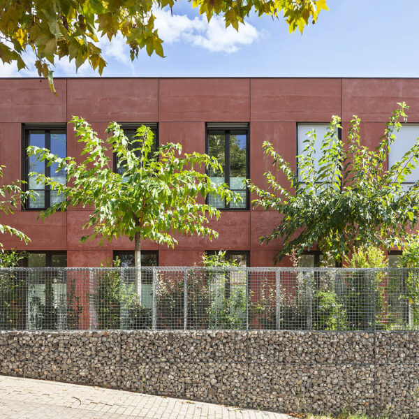 La fotografia presenta una casa passiva sostenible, dissenyada amb eficiència energètica en ment, caracteritzada per la seva façana de color terracota i finestres grans que ofereixen llum natural i ventilació adequada. L'edifici, que es troba en un ambient verd i tranquil, és envoltat per una tanca de pedra i malla metàl·lica, combinats amb vegetació que aporta un aspecte natural i sostenible. La construcció de la casa passiva en Sant Cugat és un exemple de disseny que minimitza el consum d'energia per a la calefacció i el refredar, aconseguint l'estàndard en el disseny de cases sostenibles.