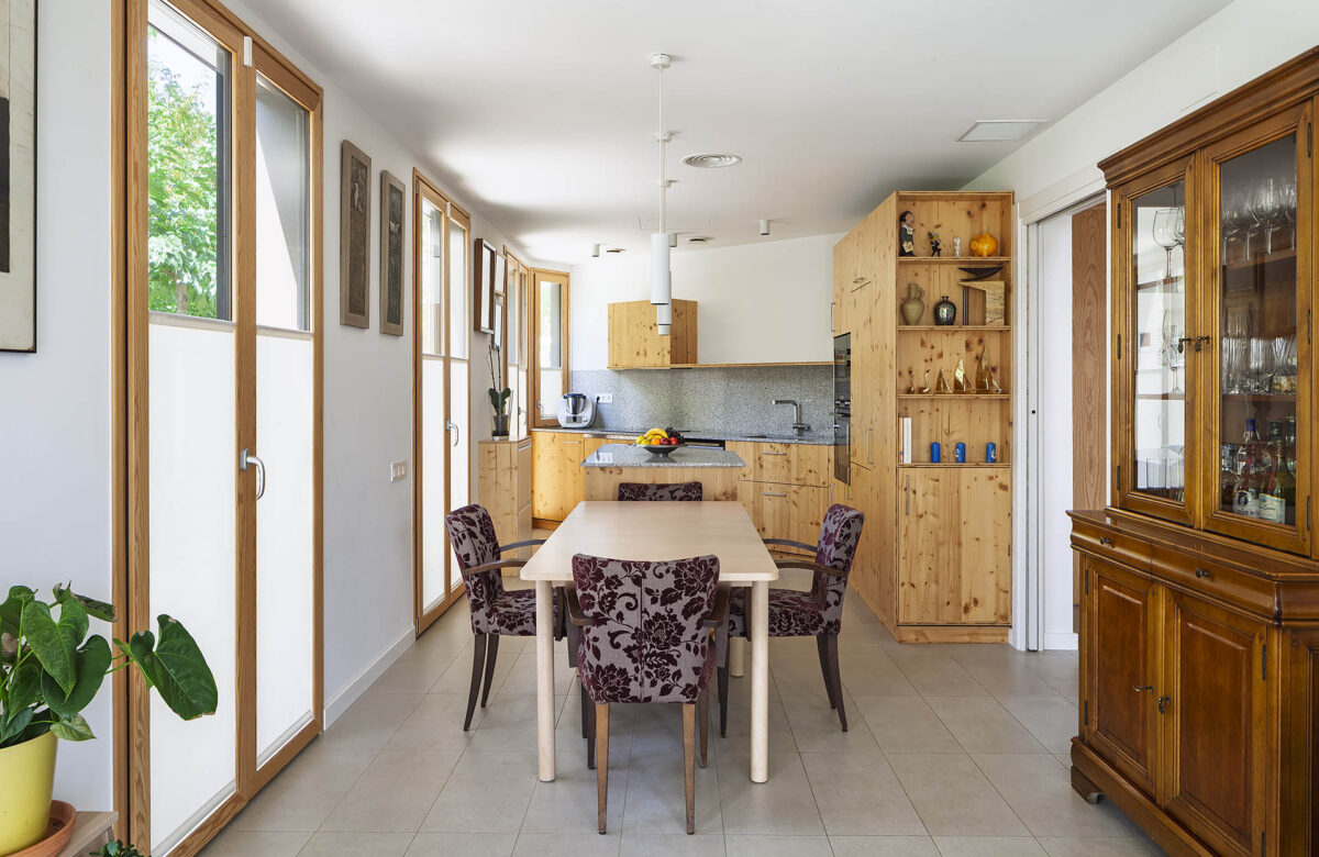 La imatge presenta un interior modern i funcional del menjador i cuina d'una casa passiva dissenyada per a maximitzar l'eficiència energètica i la sostenibilitat. L'espai de cuina i menjador, il·lumina per les finestres grans i la llum natural, i està dotat amb mobles de fusta natural i armaris de cuina, que aporten un estil de vida més sostenible i respectuós amb el medi ambient. La construcció d'una casa passiva o una casa de molt alta eficiència energètica és més que una opció, és una inversa per a un futur sostenible.